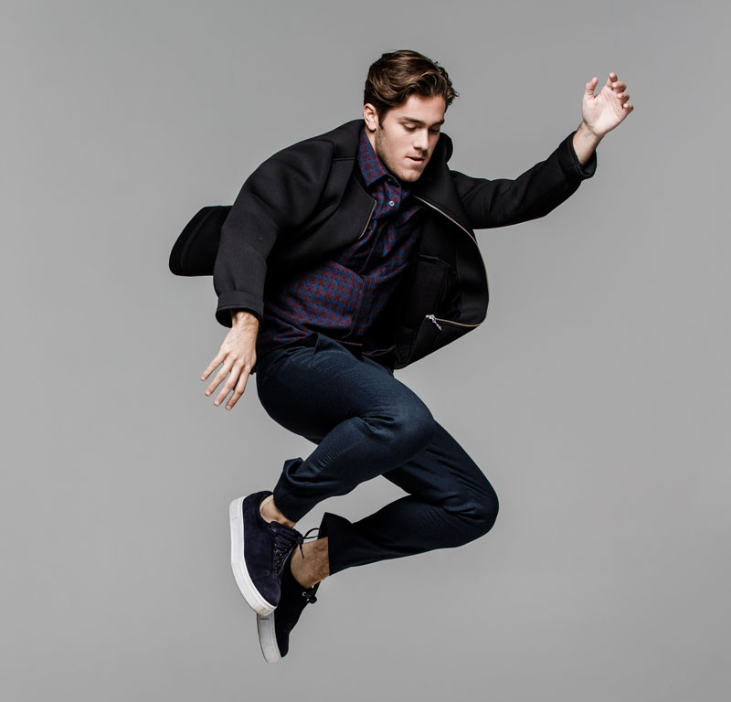 Benjamin hoppar upp i luften. Han har blå jeans, rutig skjorta och en svart jacka på sig.